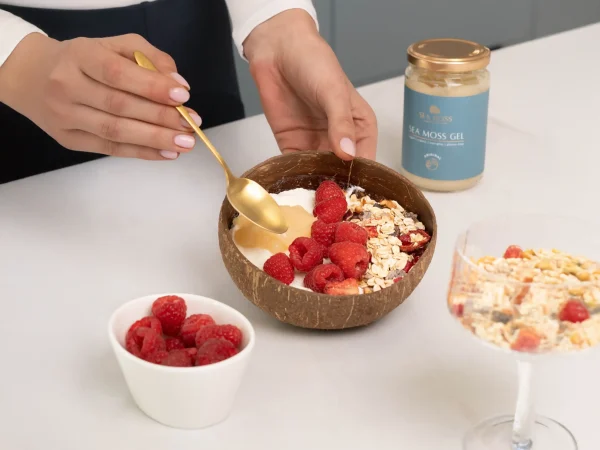 Dziewczyna przygotowuje owsiankę w misce kokosowej z malinami i żelem z mchu morskiego Sea Moss, używając złotej łyżki. Niedaleko znajduje się mała miska z malinami, a także słoik żelu z mchu morskiego i szklanka owsianki na stole.